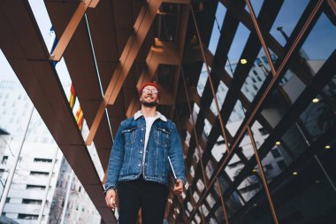 Modern şehir merkezinde yürürken elinde tablet tutan kırmızı şapkalı ve kot ceketli modern sakallı bir erkek.