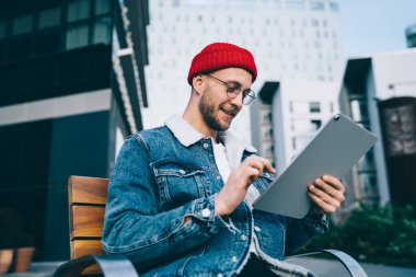 Şehrin iş merkezinde bankta otururken kırmızı şapkalı ve kot ceketli neşeli adamın tablet kullanmasından.