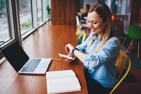 Smiling woman sitting at wooden table with new laptop device and personal planner looking at smartwatch for checking time before business meeting, optimistic female specialist in market technology