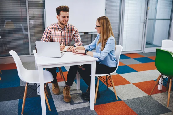 Pair of young talking people sitting at table with laptop in modern coworking space, positive female student giving private lesson on topic programming software male fellow indoors using netbook