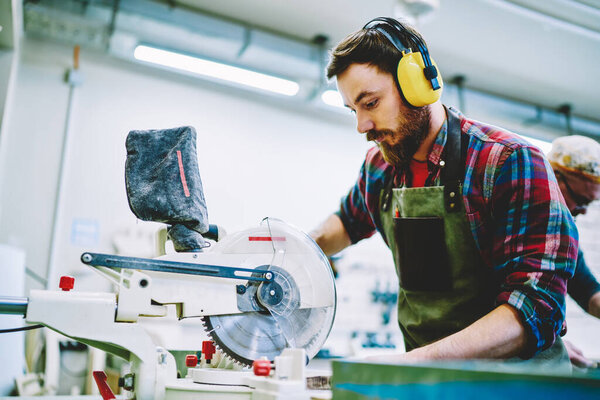 Caucasian man in headphones using circular for work with wood and timber in workshop with safety, skilled joiner using electronic equipment making details for furniture owning carpentry business