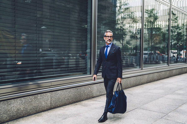 Sophisticated trendy middle aged senior man in elegant outfit carrying bag while walking confidently on street of New York city 