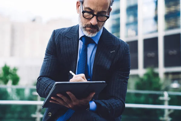 Handsome concentrated middle aged man in business suit and glasses using tablet with stylus pen while sitting against background of New York street