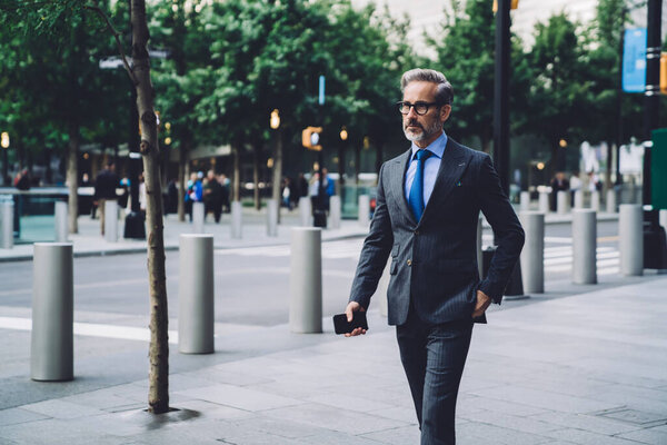 Serious middle aged man in formal clothing and glasses holding smartphone while walking in downtown area of New York City