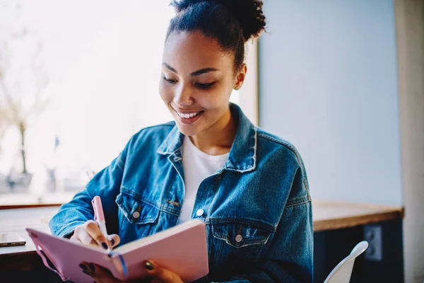 教育のための教科書で大学のコースの仕事のためのアイデアを書いてジーンズジャケットで幸せな女性学生 成功したアフリカの女性ジャーナリストは会議や将来のインタビューについての情報をメモ — ストック写真