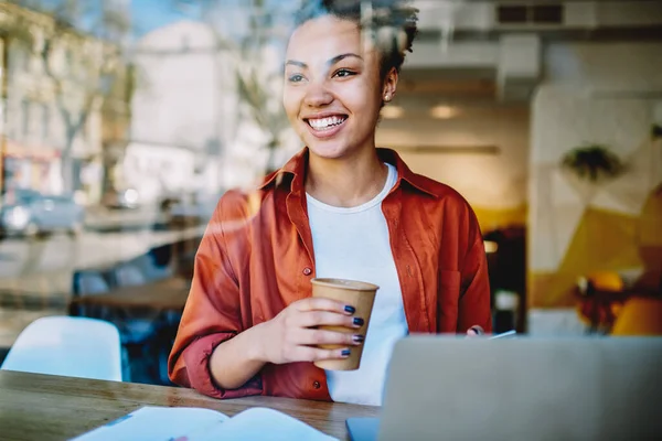 コーヒーを飲みながら紙コップを持ち キャンパス内のラップトップコンピュータとテーブルに座っている間に笑う幸せな暗い肌の女性ティーネージャー レジャーでカフェイン飲料を楽しんで興奮アフリカの女性 — ストック写真