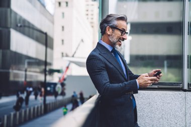 Resmi takım elbiseli ve kravatlı mutlu iş adamının New York 'ta balkonda akıllı telefondan yazı yazışının yan görüntüsü.