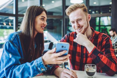 Mutlu gülümseyen adam ve kadın içerik web sitesinde cep telefonuyla komik videolar izliyor, Kafkasyalı arkadaşlar kafeteryada boş zamanlarında akıllı telefon aygıtıyla iletişim kuruyorlar.