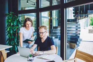 Pozitif erkek metin yazarı içerik web sitesine yazı yazarken, kadın arkadaşı elinde ders kitabıyla yanında duruyor ve dizüstü bilgisayardaki 4G bağlantısı, işbirliği konsepti hakkında danışmanlık yapıyor