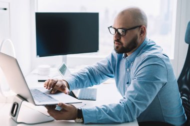 Ofis tabletinde oturan ve işe odaklanmış tabletli dizüstü bilgisayar kullanan gözlüklü kel bir adamın yan görüntüsü.