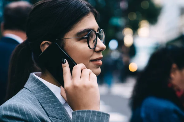 Side view of positive proud ceo in spectacles for vision correction smiling during international smartphone conversation with business partner, happy Latino manager in eyewear phoning via app