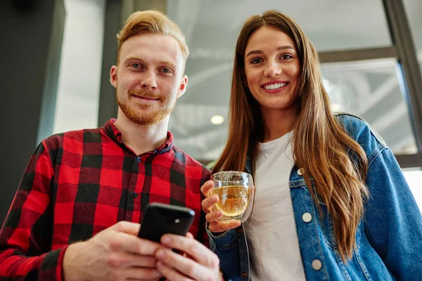 Portrait of cheerful man and woman looking at camera while enjoying fresh beverage and communication via cellphone, positive millennial users waiting for message about discounts in web store