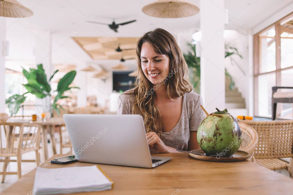 Cheerful woman in casual wear creating excited article with information about tropical fruits sitting at cafeteria in Bali and connecting to public internet, positive hipster girl enjoying chatting