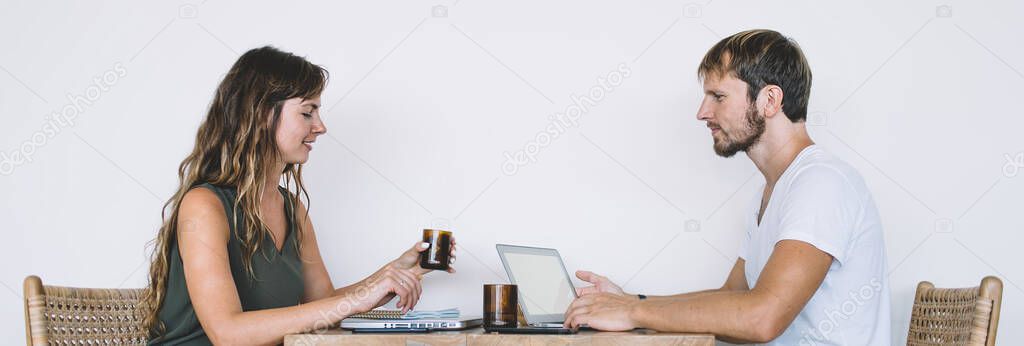 Side view of positive Caucasian male and female pair of freelancers sitting ad desktop with modern digital netbook for online working, successful digital nomads near copy space wall for advertising
