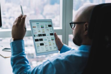 Ofiste otururken tablet kullanarak ve grafiklerin grafiklerini inceleyerek gözlüklü, kel ve bulanık bir adam.