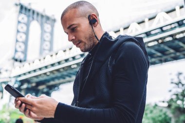 Kulaklıklı, siyah spor ceketli, bulanık şehir arka planında telefonla mesaj yazan düşünceli etnik adamın yan görüntüsü