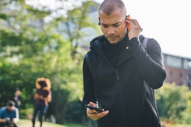 Elinde telefonla müzik seçen, arka planda bulanık kulaklıklara dokunan siyah ceketli, düşünceli genç bir erkek.