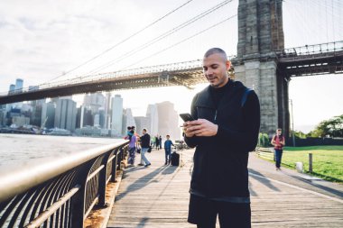 Gülümseyen atletik, traşsız, siyah spor kıyafetli genç adam şehir köprüsünün arka planında metal çitlerde akıllı telefonuyla yazı yazıyor.