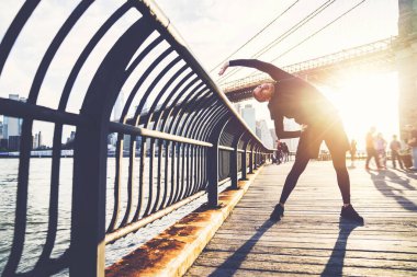 Siyah spor giyim giymiş atletik bir adam kenarda kıvrılıp şehir seti seti üzerinde ısınırken ışıl ışıl parlıyor.