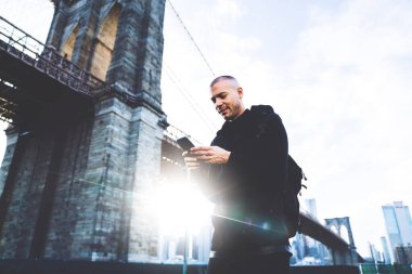 Aşağıdan neşeli, kısa saçlı, siyah spor giyim ve sırt çantalı genç adam arka plandaki devasa köprünün yakınındaki akıllı telefona dokunuyor.