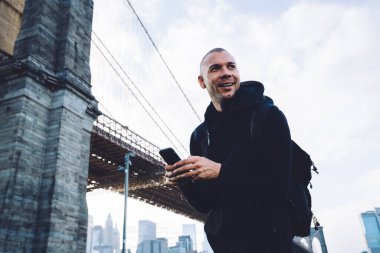 Kısa saçlı, kıllı, siyah spor giysili, sırt çantalı genç bir adam. Gündüz vakti köprüde akıllı telefondan yazı yazıyor ve başka yere bakıyor.