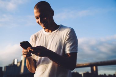 Genç gülümseyen kısa saçlı adam gölgede duruyor ve akıllı telefonda daktilo yazıyor. 