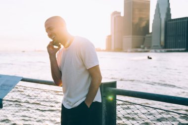 Günlük giysiler içinde suyun yanında duran korkuluklara yaslanan ve telefonda konuşan su manzaralı genç adam. 