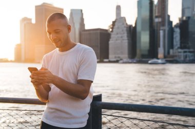 Beyaz tişörtlü genç adam güneş ışığında duruyor. Suyun yanında eskrime yaslanıyor ve telefonda gülümsüyor.