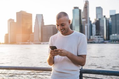 Beyaz tişörtlü, kısa saçlı, güneş ışığında duran, şehrin sularında eskrim yaparken yaslanan ve akıllı telefondan yazarken eğlenen genç bir adam.