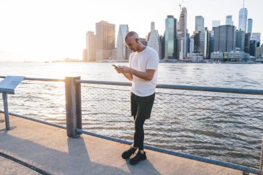 Kulaklıklı yakışıklı, kel bir adam modern dijital aletlerle interneti tarıyor ve ciddi bir şekilde aşağı bakıyor ve şehir arka planında nehrin yanında duruyor.