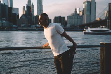 Yetişkin yakışıklı atletik, spor kıyafetlerine odaklanmış, ısınan bir erkek, şehir arka planında, gökdelenler ve teknelerle nehir kenarındaki kulaklıklara bakıyor.