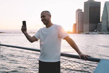 Genç, gülümseyen, kulaklıklı, akıllı telefonuyla selfie çeken ve nehir ve şehrin zincire bağlı metal çitlere yaslanan yakışıklı bir etnik adam.