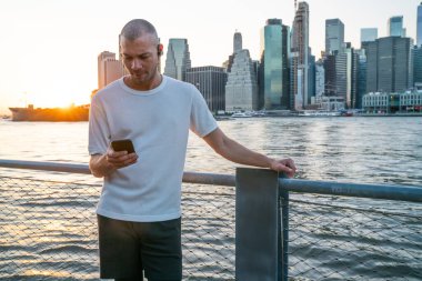 Kulaklıklı, akıllı telefon kullanan ve şehrin arka planında metalik çit ve nehrin yanında duran beyaz tişörtlü genç bir etnik adam.