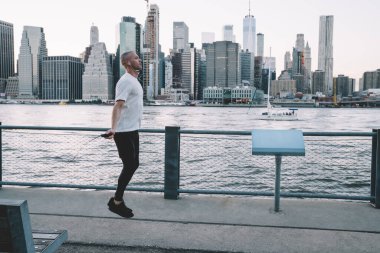 Doğu Nehri ve New York 'un arka planına karşı metal çitlerin yanında ip atlarken kısa saçlı, kulaklıkla müzik dinleyen genç bir adamın yan görüntüsü.