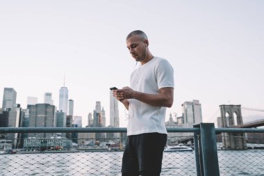 Spor giyinen, kulaklıkla müzik dinleyen ve nehirde yürürken akıllı telefon kullanan genç bir adamın yan görüntüsü. 