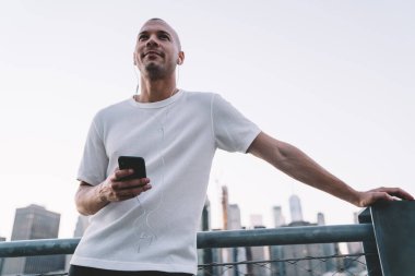 Beyaz tişörtlü, gülümseyen, kulaklıkla müzik dinleyen, akıllı telefon tutan ve modern şehrin arka planına dayanan genç bir adamdan. 