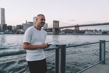 Beyaz gömlekli, kulaklıklı, akıllı telefonlu, metalik çitlere ve nehre yaslanmış genç, gülümseyen kısa saçlı bir adam. 