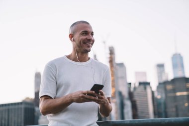 Gülümseyen, kısa saçlı, kısa saçlı, kulaklıklı ve akıllı telefonlu yakışıklı bir adam çitin yanında dikilirken gözlerini kaçırıyor. 