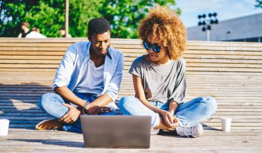 Düşünceli Afro-Amerikalı arkadaşlar modern dizüstü bilgisayarda internet üzerinden şehir ortamında kablosuz internet kullanıyorlar. Koyu tenli genç adam ve kadın netbook ile ahşap bankta oturuyor.