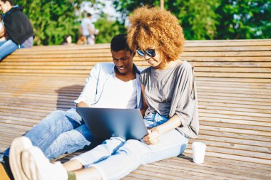 Gündelik giyinmiş Afro-Amerikan çifti şehir ortamında ahşap bankta dinleniyor ve laptopta uygulama kuruyor. İnternette özel ders izleyen koyu tenli iki arkadaş.