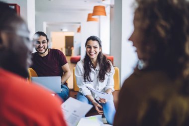 Çeşitli grup arkadaşları olan neşeli genç kadın ve erkek yeni projeyi tartışırken ellerinde kağıtlar ve laptopla salonda oturuyorlar.