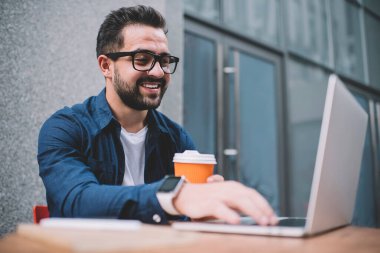 Neşeli adam metin yazarı web sitesinde bilgi yayınlamak için yazı yazıyor şehir sokak kafeteryasında uzaktan çalışıyor, olumlu erkek programcı internetteki webinar için ayrılmış biletten mutlu.
