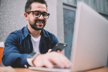 Mobil uygulama ile çalışan mutlu erkek yazılım geliştirici modern laptop aygıtı ile iletişim için kamu sistemi yaratıyor, cep telefonundan Netbook 'a içerik metni yazan göz giysisi içindeki genç adam