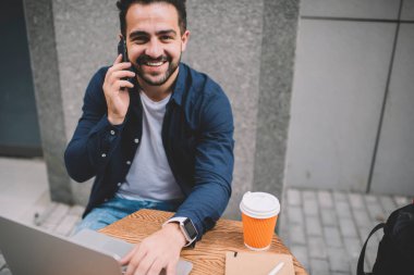 Kameraya bakan neşeli erkek programcının portresi sohbet etmek için arkadaşına seslenirken, sokak kafeteryasında oturan ve halka açık interneti uzaktan çalışmak ve telefon etmek için kullanan yetenekli bir adam.