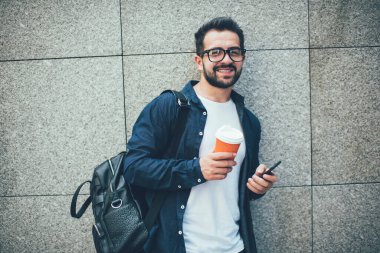 Şehir duvarının yanında şık bir sırt çantası olan ve kameraya gülümseyen neşeli, başarılı erkek turistin yarım boy portresi. Seyahat kaçamağının keyfini çıkarıyor.