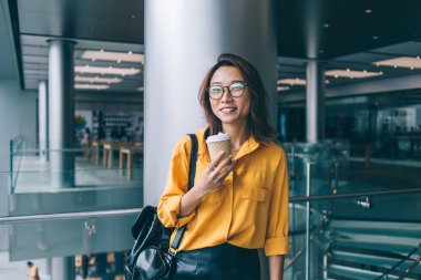 Hong Kong 'da alışveriş merkezinde gülümseyen kameraya bakan, elinde bir kutu kahve ve çantayla mutlu, ince Asyalı bir kadın.