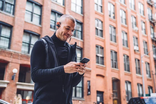 Delighted bald bristly athletic man in headphones and in black sportswear tapping on mobile in daylight on urban blurred background