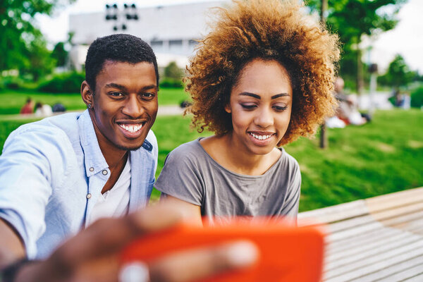 Cheerful african american young man and woman laughing while making selfie photos on modern smartphone device for blog.Positive couple in love taking picture on front camera of digital telephone