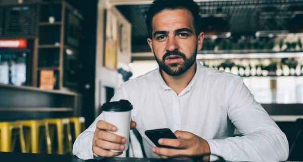 Portrait of experienced male employee in casual white shirt posing for camera while checking digital marketing on financial website using high speed internet connection in public coffee shop