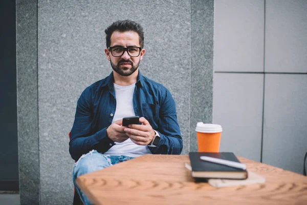 Portrait of intelligent male student sitting at street coffee shop and looking at camera while searching knowledge information on website, millennial man in classic eyeglasses using 4g on cellphone